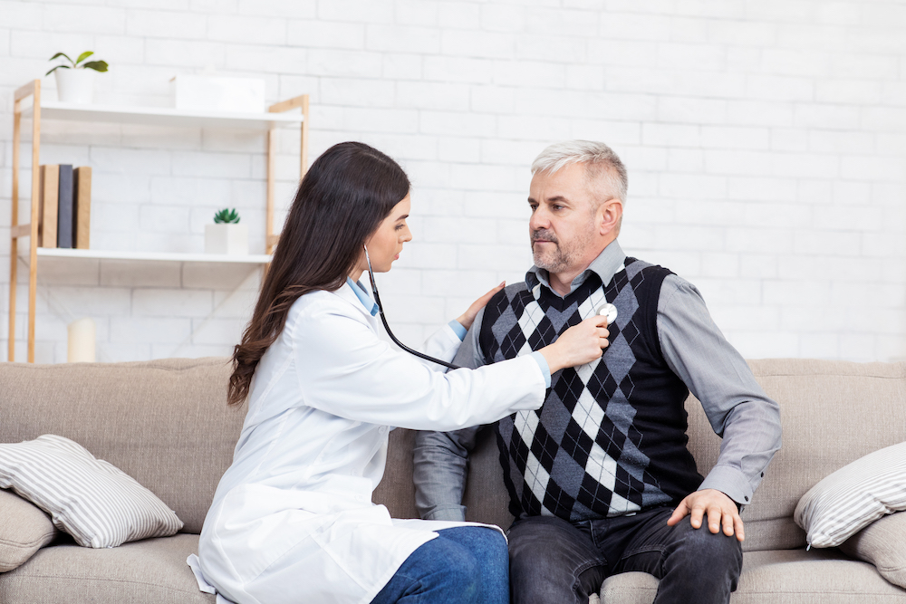 Protection of health of senior patient and risk group, colds and heart disease. Young woman doctor examines patient and listens to breathing with stethoscope, man sits on sofa in office or home