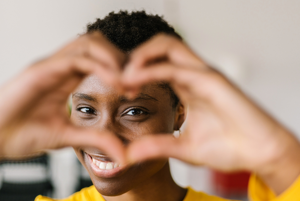 Portrait of the young smiling lovely multiracial woman showing shape heart with hands and looking through it with toothy smile while posing at her home