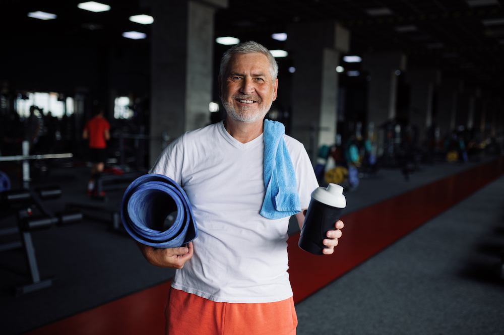 Happy old man with bottle of water, towel and mat, gym interior on background. Sportive grandpa on fitness training in sport centre. Healthy lifestyle, health care, elderly sportsman. improve your prostate health