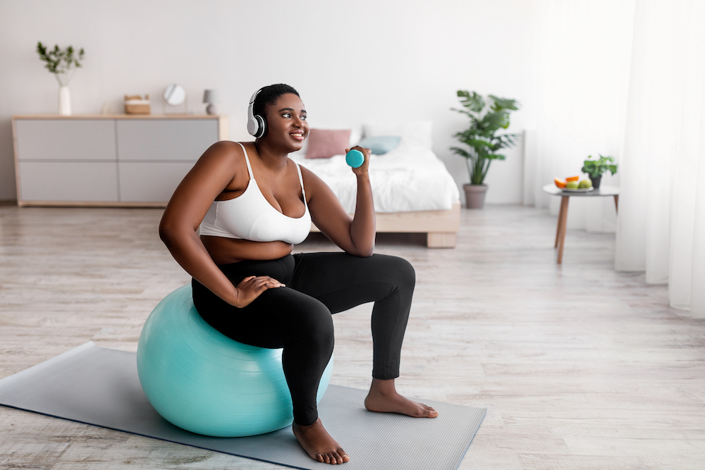 Overweight Afro woman exercising with dumbbells, sitting on fitball, listening to music in headphones indoors, copy space. Curvy black lady doing home fitness, losing weight, full length