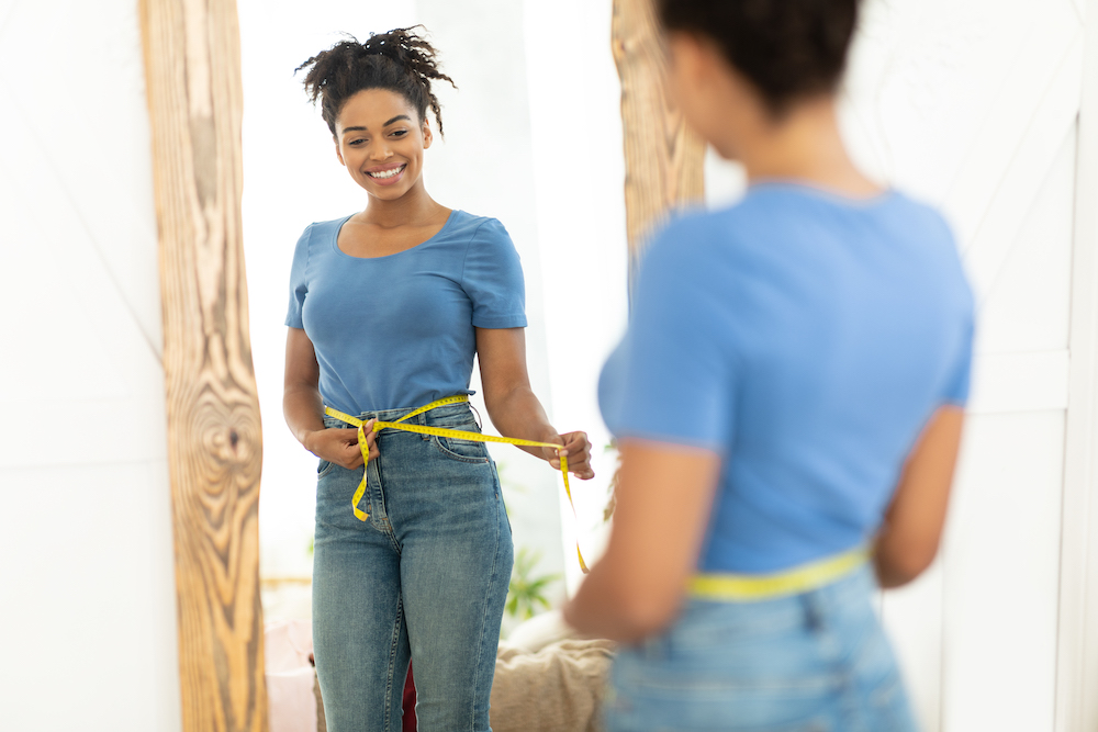Happy Black Girl After Weight Loss Measuring Thin Waist With Tape Standing Near Mirror At Home. Slimming Concept. Saxenda