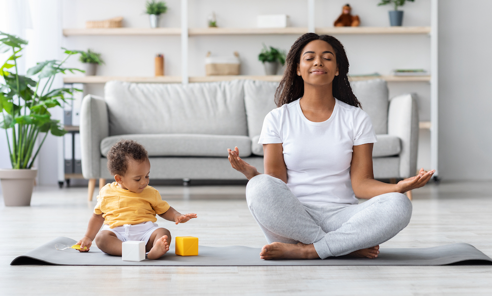 Mental Wellness Concept. Calm Happy Black Mother Meditating With Baby At Home, African Lady Sitting In Lotus Position With Closed Eyes, Practicing Yoga In Living Room With Child Playing Around